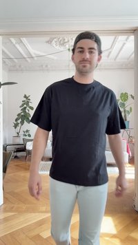 T-shirt nera a maniche corte in cotone, vestibilità comoda, abbinata a jeans azzurri chiari. Ambientazione interna con pavimento in legno e piante visibili.