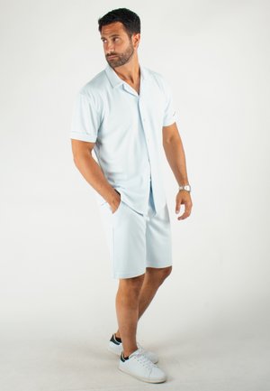 Chemise à manches courtes bleu clair à boutons associée à un short assorti, le tout en matériau lisse et respirant ; le mannequin porte des baskets blanches.