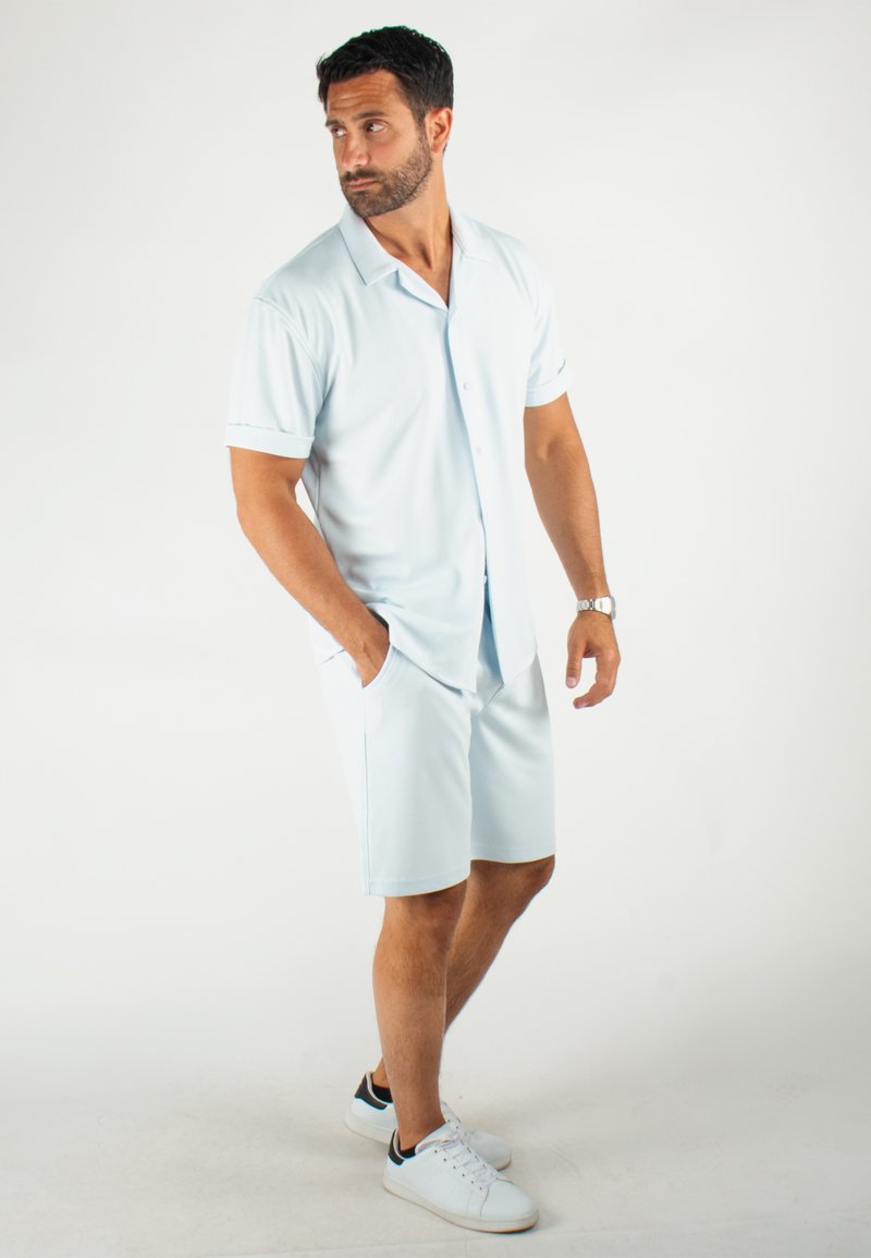 Chemise à manches courtes bleu clair à boutons associée à un short assorti, le tout en matériau lisse et respirant ; le mannequin porte des baskets blanches.