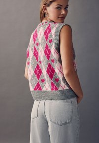 Sleeveless knit vest featuring a pink, gray, and white argyle pattern with heart designs, ribbed gray hem, and button accents at the back.