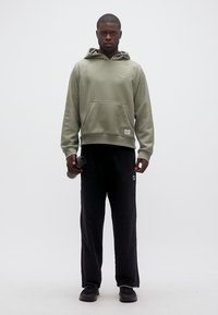 Άνδρας όρθιος που φορά γκρι φούτερ με κουκούλα, μαύρο φόρμας και μαύρα αθλητικά παπούτσια, κρατώντας ένα μαύρο παγούρι νερού στο δεξί του χέρι.