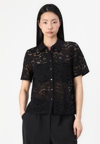 Chemise en dentelle noire à manches courtes, fermeture à boutons, motif floral et texture douce. Associée à un pantalon noir.