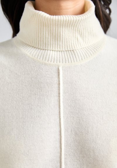 Sweter w kolorze kremowym z golfem, z prążkowanym kołnierzem, gładką fakturą oraz pionowym szwem biegnącym przez środek przodu.