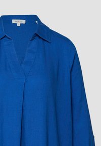 Blouse bleue à manches longues avec col en V et col, comportant une petite étiquette de marque à l'intérieur de l'encolure.