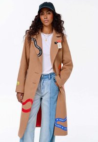 Žena s kudrnatými vlasy má na sobě dlouhý béžový kabát s barevnými nášivkami "DON'T STOP", bílým tričkem, světlomodrými džínami, černou kšiltovkou a vrstvenými náhrdelníky.