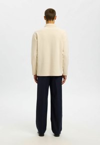 Bež majica dugih rukava s rebrastim uzorkom i klasičnom kragnom, u kombinaciji s širokim mornarsko plavim hlačama. Stražnja strana prikazuje bešavne šavove i opušteni kroj.