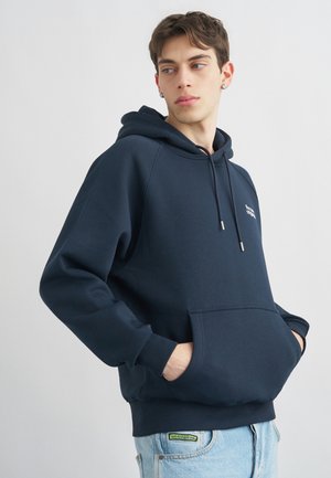 Noor mees lühikeste juustega, kes kannab tumedat mereväe sinist kapuutsi ja hoiab käed esitaskus, vaadates küljele heleda ühevärvilise tausta ees.