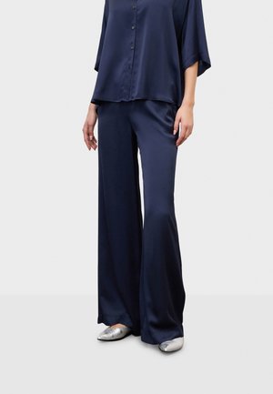 Femme portant un pantalon en satin bleu foncé ample et une chemise assortie à boutons, associée à des ballerines pointues argentées, debout sur un fond clair.