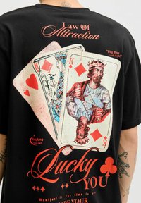 Zwart t-shirt met grafische print van speelkaarten, rode textelementen en vintage afbeeldingen van een koning in wapenrusting.