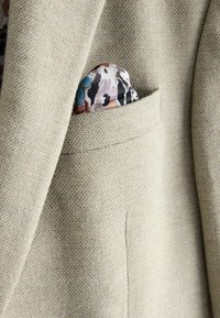 Blazer texturizado beige con un bolsillo en el pecho que exhibe un pañuelo de bolsillo con un patrón en tonos de crema, marrón y teal.