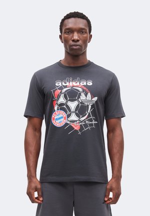 Muž nosiaci tmavé tričko Adidas s futbalovou loptou, logom FC Bayern Mníchov a textom „Bayern München“ vpredu, stojaci na jednoduchom pozadí.