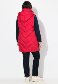 Rote ärmellose Steppweste mit Kapuze, die ein Chevron-Muster und Reißverschlusstaschen aufweist, getragen über einem dunklen Langarmshirt und dunklen Jeans.