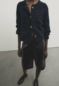 Marineblauer strukturierter Cardigan mit goldenen Knöpfen, zwei Vorderaschen und lockerem Schnitt. Kombiniert mit glänzenden schwarzen Lederhosen und Loafern.