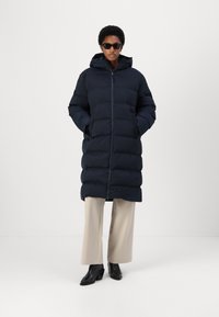 Navy puffer coat with a hood, zipper closure, and side pockets; quilted design with a smooth finish. Paired with beige wide-leg pants and black boots.