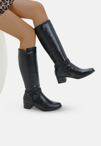 Botas negras hasta la rodilla hechas de cuero suave, con un tacón bajo, un zipper lateral y correas decorativas con acentos metálicos.
