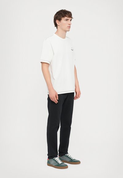 White cotton t-shirt with a smiley face design, paired with black pants and green sneakers featuring white laces and brown rubber soles.