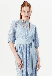 Lacekjole i lyseblå med pufferm, høy hals, knappelukking og matchende midjebelte, med intrikate blomsterdesign.