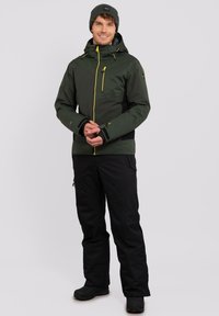 Giacca impermeabile verde con zip gialle, dettagli neri, cappuccio regolabile e pantaloni neri. Il modello indossa un berretto abbinato e stivali.