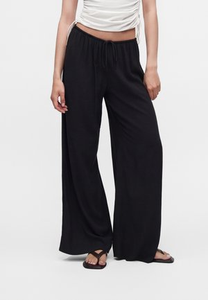 Kvinde iført sorte bukser med vide ben og elastisk talje samt hvid rynket cropped top, stående med fødderne i sorte flip-flops.