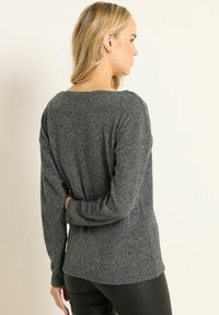 Frau mit blonden Haaren, die einen dunkelgrauen, langärmligen Strickpullover und schwarze Hose trägt, steht mit einem Arm hinter ihrem Rücken.