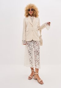 Blazer beige su misura con due bottoni, maniche in pizzo e abito maxi in pizzo sotto. Tacchi beige con cinturini e occhiali da sole rotondi completano il look.