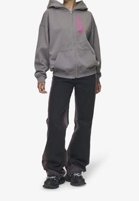 Felpa grigia con zip e cappuccio, con testo rosa sul lato sinistro. Abbinata a jeans neri a gamba larga e sneakers nere.