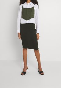 Olivgrön croppad topp med tunna axelband över en vit skjorta med knappar, tillsammans med en mörkgrön figurnära knälång kjol och svarta högklackade mule-skor.