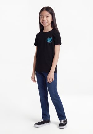 Jong meisje met lang donker haar, gekleed in een zwart Santa Cruz t-shirt, blauwe jeans en zwarte sneakers, staand en glimlachend tegen een witte achtergrond.