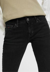 Jeans de denim negro con un corte slim, que cuentan con un diseño de cinco bolsillos, sutiles detalles de costura y un cierre de botón metálico. Parte superior de punto texturizado visible.