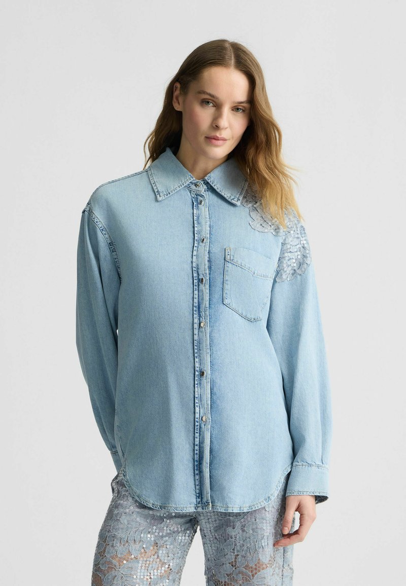 Camicia di denim blu chiaro con colletto a punta, chiusura frontale con bottoni e tasca. Presenta dettagli decorativi in paillettes su una spalla.