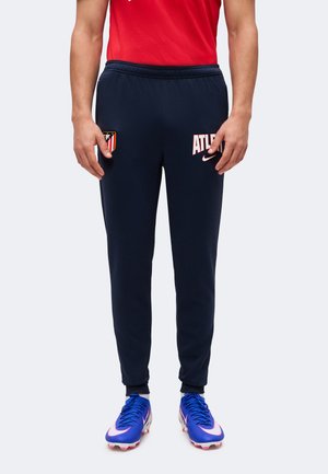 Homme portant un pantalon de sport bleu marine avec le logo de l'Atletico Madrid et des chaussures de football Nike bleues.