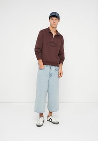 Czerwono-brązowy rozpinany sweter z napisem "ORIGINALS", jasnoniebieskie szerokie dżinsy i białe/szare sneakersy. Czapka z niebieskiego denimu.