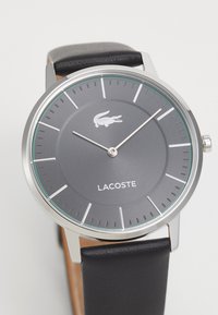 Rund grå klokke med sølvdetaljer, svart lærrem og Lacoste-logo på urskiven. Minimalistisk design med klare timemarkører.