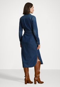 Abito chemisier in denim blu scuro, maniche lunghe, vita cinturata, spacchi laterali, abbinato a stivali al ginocchio con tacco marroni che completano l'outfit.