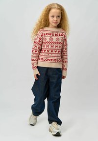 Pulover tricotat bej cu modele festive roșii care includ inimi și panglici, asortat cu pantaloni largi din denim întunecat și adidași deschiși.