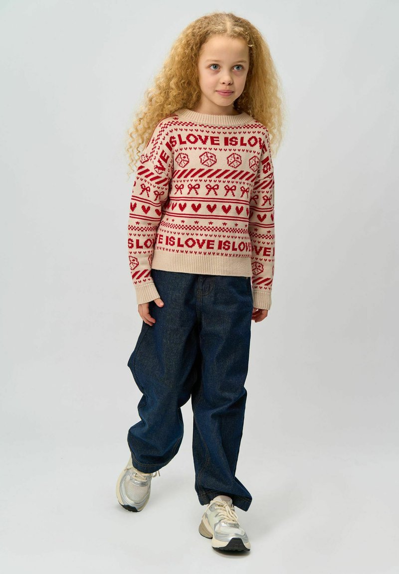 Pulover tricotat bej cu modele festive roșii care includ inimi și panglici, asortat cu pantaloni largi din denim întunecat și adidași deschiși.