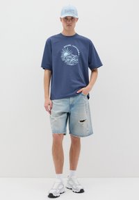 Camiseta de algodón azul marino con un gráfico claro de un sol y olas. Pantalones cortos de mezclilla ligera con un efecto desgastado y zapatillas deportivas blancas. Gorra azul clara.