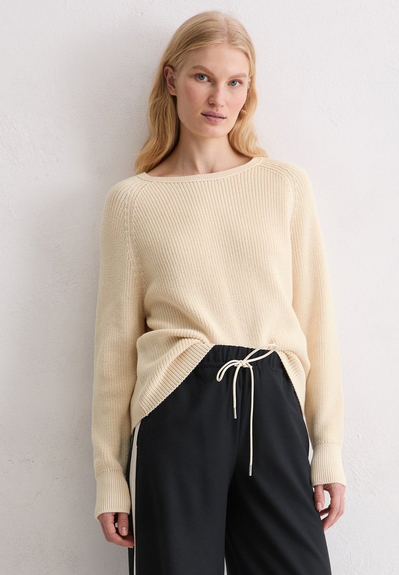 Beige geribbelde gebreide trui met een wijde halslijn en verlaagde schouders, gecombineerd met zwarte broek met trekkoord en witte zijstreep.