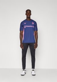Maillot de sport marine à manches courtes, avec des accents roses et un logo, assorti à un pantalon noir ajusté et des chaussures de sport blanches.