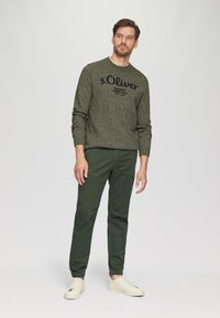 Donkergroene gebreide trui met zwarte tekst, gecombineerd met groene taps toelopende broek en lichte sneakers. De stof is gestructureerd en comfortabel.