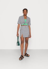 Top cropped em gingham preto e branco amarrado à frente, combinado com uma saia correspondente com uma abertura lateral. Os acessórios incluem uma mala verde e sandálias pretas.