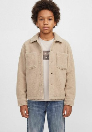 Un jeune homme porte une veste beige en sherpa à deux poches poitrine sur un tee-shirt blanc à motif graphique et un jean bleu.
