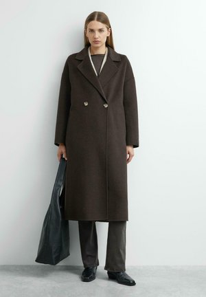 Kobieta w długim ciemnobrązowym płaszczu dwurzędowym, czarnych spodniach, czarnych butach, trzymająca dużą czarną torbę typu tote na tle białej ściany.
