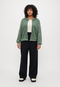 Veste zippée perforée verte avec des poignets larges, portée sur un haut crème, associée à un pantalon large noir et des chaussures de couleur claire.