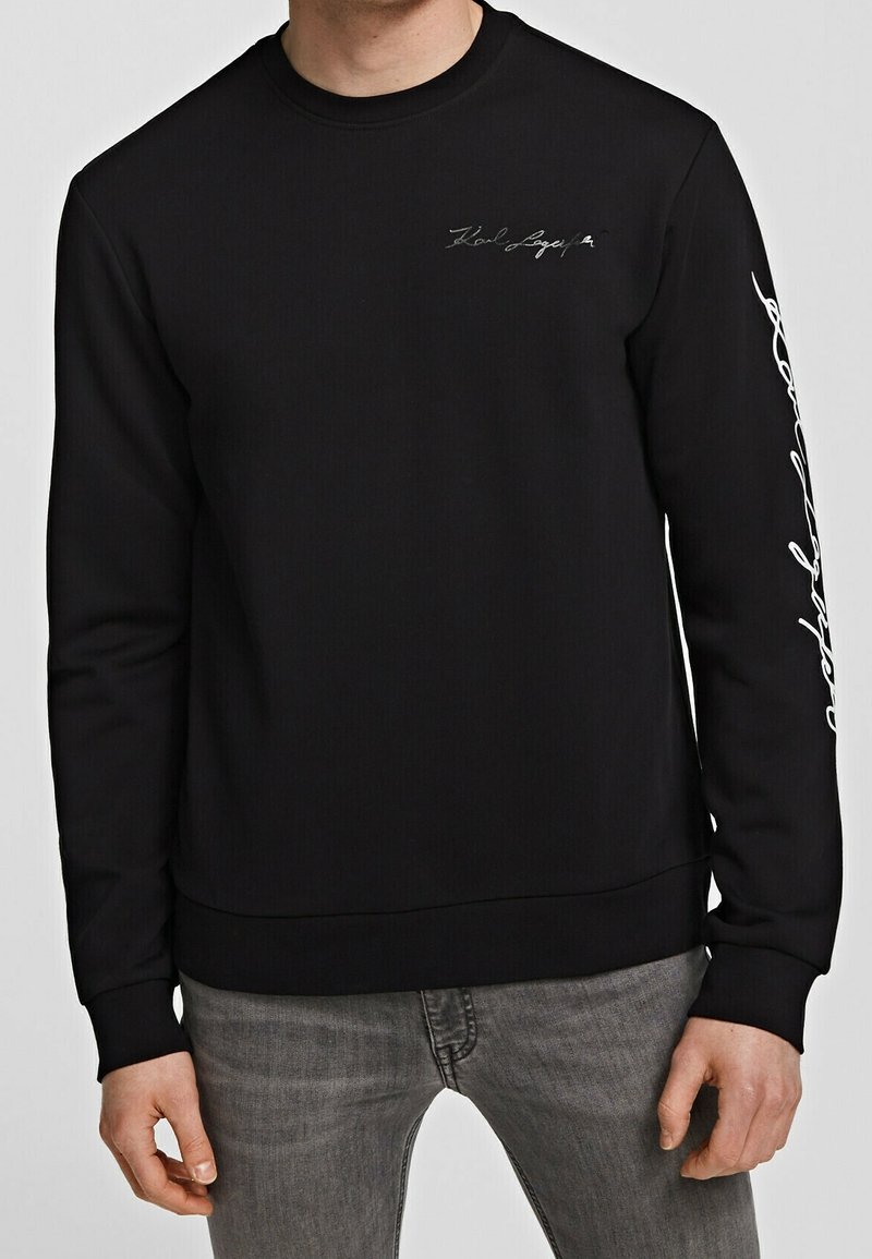 Hombre con sudadera de manga larga negra y un pequeño logotipo en cursiva en el pecho, con una escritura blanca que baja por la manga izquierda, combinada con jeans grises.