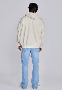 Beiger Kapuzenpullover mit überschnittenen Schultern, kombiniert mit hellblauen Jeans. Der Pullover hat eine lockere Passform und gerippte Bündchen.