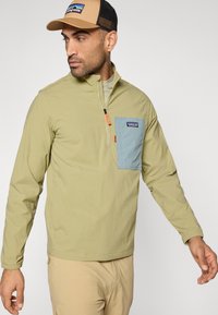 Ανοιχτό πράσινο πουλόβερ με φερμουάρ στο ένα τέταρτο, μπλε τσέπη στο στήθος, με ελαστικά μανίκια και σήμα λογότυπου. Λείο ύφασμα, casual σχέδιο.