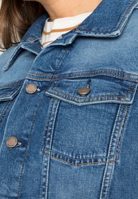 Veste en denim bleu moyen, présentant un design classique avec poches poitrine, quincaillerie en bronze et coutures orange contrastantes. Tissu texturé.