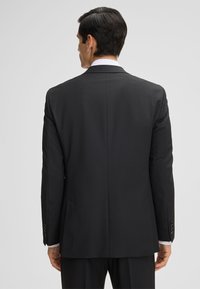 Giacca nera su misura, con taglio aderente, apertura singola sul retro, texture liscia, revers a punta e polsini con bottoni.