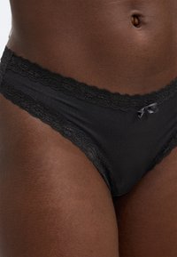 Μαύρα μπικίνι εσώρουχα με δαντέλα, τελείωμα σατέν, με μικρό φιόγκο μπροστά και ομαλή υφή σε όλη την επιφάνεια.
